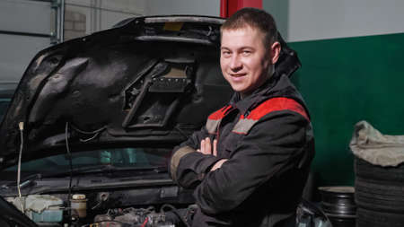 Cheerful technician with crossed arms in grey uniform stands near renovated automobile with open bonnet in workshop of car service stationの写真素材