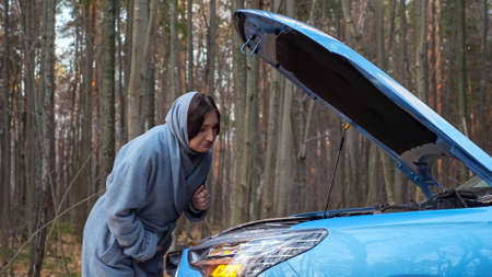 Young woman in coat and scarf on head looks under hood of broken car with alarm headlights on roadside near autumn forest with bare trees, side viewの写真素材