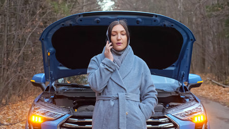 Woman driver in coat and scarf stands against broken car with open hood and calls emergency service...