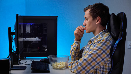 Young brunet man in checkered shirt smiles watching stream with interest on large computer screen...