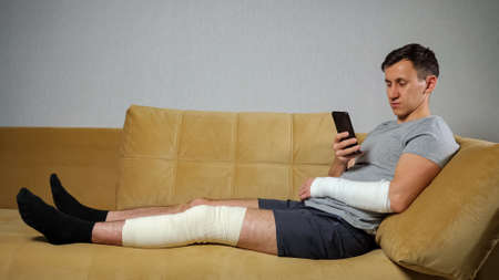 Brunet man in grey T-shirt with injured knee in bandage and broken forearm scrolls through social nets using smartphone sitting on sofa.の写真素材