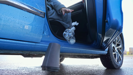 Woman opens foreign car door and throws away plastic bottle and coffee cup onto asphalt wet from...