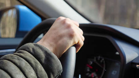 Man in dark warm sheepskin coat holds steering wheel confidently and drives foreign car along road between rows of trees in autumn forest closeupの写真素材