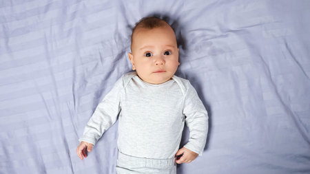 Little infant boy with short blond hair wearing white cotton bodysuit and diaper lies on comfortable...