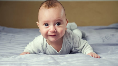 Baby boy with short blond hair in white cotton bodysuit lies on tummy and tries to crawl on...