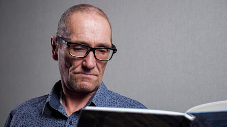 Focused senior man in dark blue shirt and elegant black-rimmed glasses for bad eyesight reads large print paper book against grey wall closeup.の写真素材