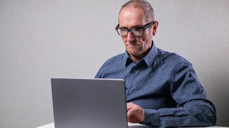 Senior man in classic dark blue dotted shirt with black-rimmed glasses works in computer sitting at white desk in office against grey wall.の写真素材