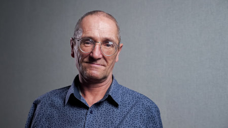 Elderly man with high forehead in classic blue shirt with bad eyesight wears elegant stylish round shaped glasses smiling at grey wall closeup.の写真素材
