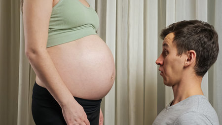 Surprised brunet father looks at pregnant belly and listens to baby moves in tummy of wife wearing...