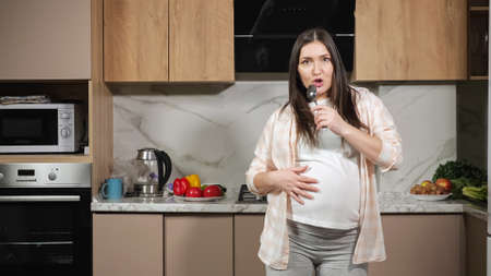 Pregnant woman with long brunette hair in comfortable domestic clothes dances and sings with spoon imitating microphone in kitchen near counters.の写真素材
