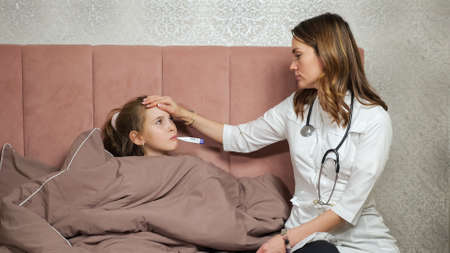 Lady doctor in white robe measures temperature putting thermometer into mouth of sick teen girl lying on bed covered with blanket in bedroom at home.の写真素材