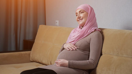Muslim woman in pink-colored hijab and warm dress caresses gently pregnant belly and smiles happily sitting on comfortable sofa in living room.の写真素材