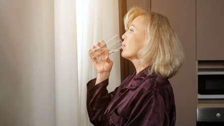 Middle aged blonde long-haired woman with bangs drinks glass of water for health enjoying morning view outside window standing in kitchen, sunlight.の写真素材