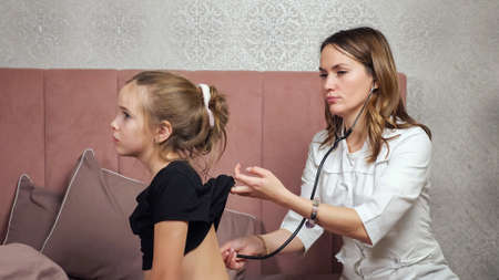 Doctor woman in white coat listens to lungs of sick teen girl with stethoscope sitting on bed against patterned wall in bedroom at home.の写真素材
