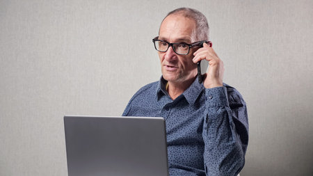 Elderly man with black-rimmed glasses in classic blue dotted shirt talks on mobile phone working on...