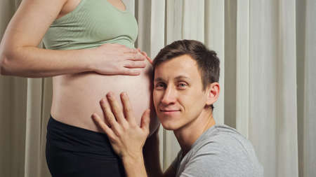 Brunet young husband kisses and listens to unborn baby moves in stomach of pregnant wife wearing sports top and leggings in living room, closeup.の写真素材