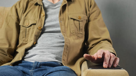 Man in mustard jacket sits in soft armchair tapping fingers on armrest against grey wall at home....