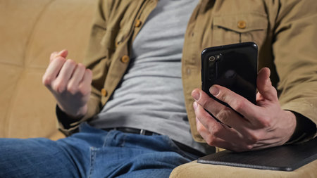 Man in mustard jacket wins lottery checking result in mobile phone at home. Player enjoys online...