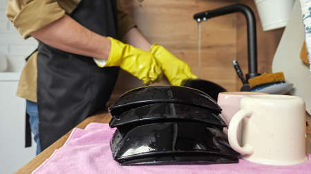 Man washes black plates under flowing water from faucet stacking on dry towel on countertop. Householder in apron does chores in kitchen closeupの写真素材