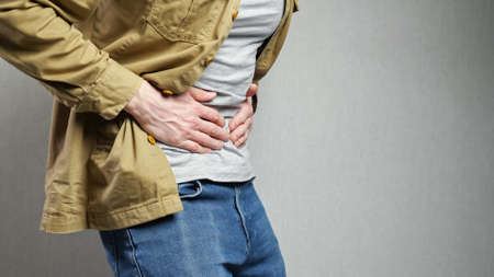 Man suffers from sudden severe pain in stomach touching belly with hands. Male person has abdominal health problems standing near grey wall, side viewの写真素材