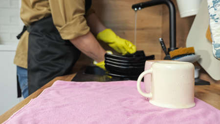 Man washes black plates under flowing water from faucet stacking on dry towel on countertop. Householder in apron does chores in kitchen, closeupの写真素材