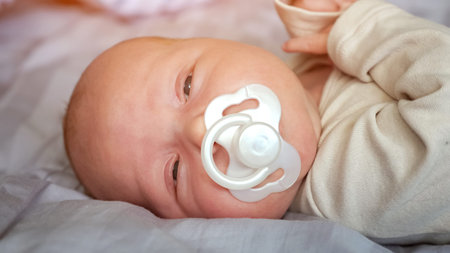 Adorable newborn baby girl sucks pacifier lying in bed at home. Calm little kid in beige bodysuit...
