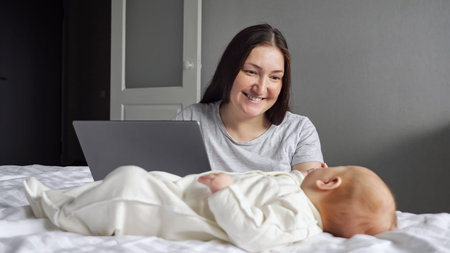 Mother pays attention to little baby girl sitting near bed and working on laptop. Newborn child...