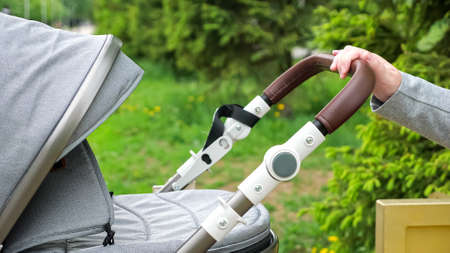 Mother hand rocks stroller with baby against blurred green trees and bushes. Woman sits on bench in park breathing fresh air closeupの写真素材