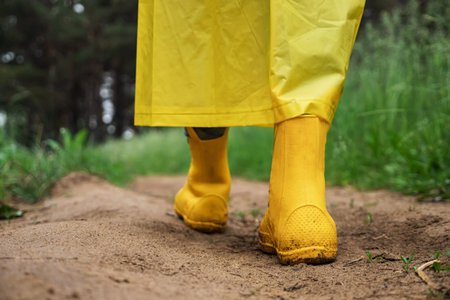 Person legs walk on wet road in rainy weather in green forest. Child dressed in yellow rain-boots and raincoat. Grass grows around path closeupの写真素材