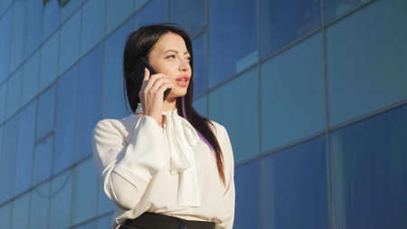 Young lady manager stands against business office and talks with partner on phone. Woman with plump lips dressed in white blouse with bow communicatesの写真素材