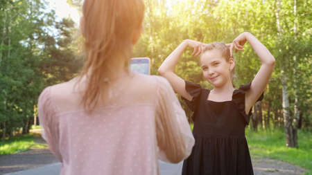 Couple of teen girls makes video vlog on phone camera. Schoolgirl wearing black dress poses grimacing in park. Teenager wearing pink dress records video, sunlightの写真素材