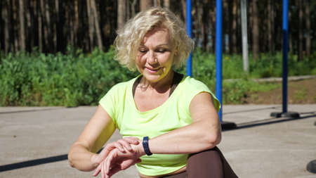 Mature woman checks pulse on fitness bracelet after powerful workout. Young blonde pensioner rests sitting on asphalt sports ground closeupの写真素材