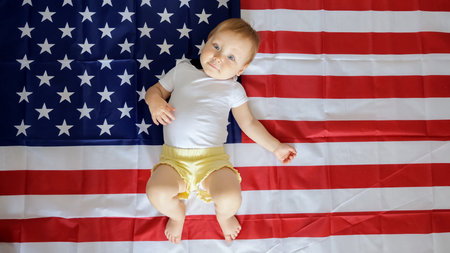 Cheerful toddler with blonde hair lies on flag of USA in light studio. Resting child with attraction...