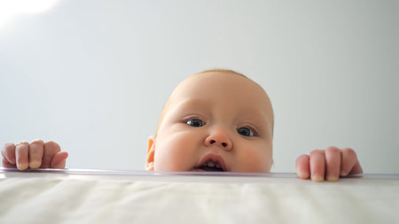 Baby girl pulls up hands looking from under white table. Small daughter peeks out from beneath table...