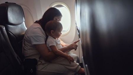 Mother caresses and playfully engages with toddler girl. Mom holds child securely in arms as family soars through skies on journey by planeの写真素材