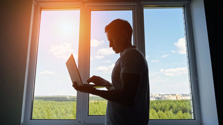 Male office worker standing to window scrolls social media on smartphone. Man employee in t-shirt...