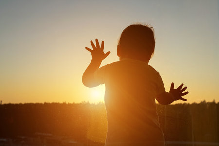 Silhouette of child stands against backdrop of window. Vivid hues of setting sun visible through glass creates captivating and serene scene, copyspaceの写真素材