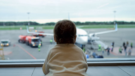 Toddler standing by large window looks at people getting aboard plane. Girl follows eyes for passengers preparing to embark on journeyの写真素材