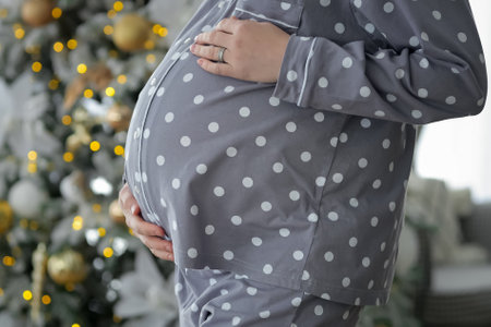 Pregnant woman strokes future baby in belly waiting for child. Lady wearing grey pyjama gets ready to celebrate Christmas near decorated tree at homeの写真素材
