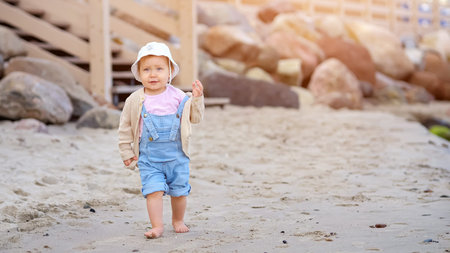 Funny toddler girl smiles running barefoot on sandy beach adjusting blouse on background of large blocks of stones and wooden stairs, sunlightの写真素材