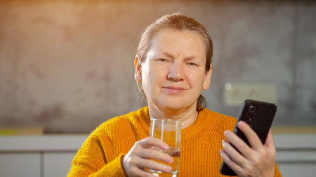 Senior woman in cozy sweater reading news on phone drinks glass of water. Lady hydrating scrolls...