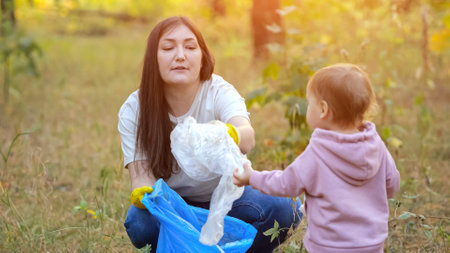 Toddler girl contributes to environmental conservation by helping mom in cleaning up litter. Child helps mother to pick up garbage, sunlightの写真素材