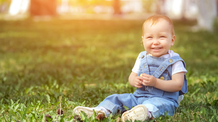 Girl toddler in denim pantsuit laughing and smiling in park. Small kid sits on green grass lawn...