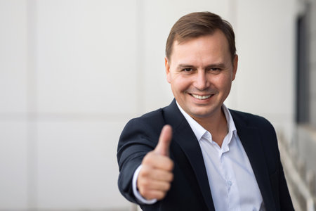 Confident adult businessman in formal suit stands near office and smiles raising thumb-up. Mature office worker expresses successful deal end, copyspaceの写真素材