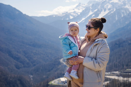 Mother with hair-bun looking at toddler daughter in arms against sunlit mountain with snowy peaks. Girl looks around with confused expressionの写真素材