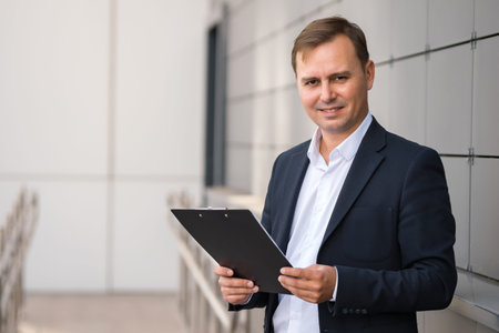 Confident mature businessman in suit stays outdoors and smiles in front of camera holding documents in hands. Office worker clearly satisfied by dealの写真素材