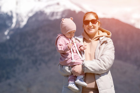 Smiling mother in black sunglasses holding daughter enjoys visiting mountain resort with snowy...
