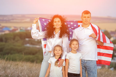 Patriotic family holds USA flag standing against rural area with residential buildings. Parents with children pose for photoshoot on field, sunlightの写真素材