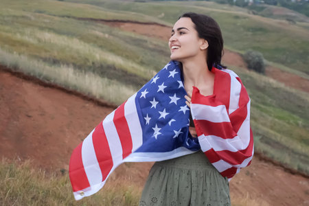 Cheerful female wraps in USA flag standing on hilly place with picturesque view. Concept of love for...