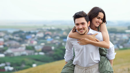 Happy bearded man runs carrying girlfriend on back up hill overlooking large village in cloudy weather. Have fun and spend time together, copyspaceの写真素材